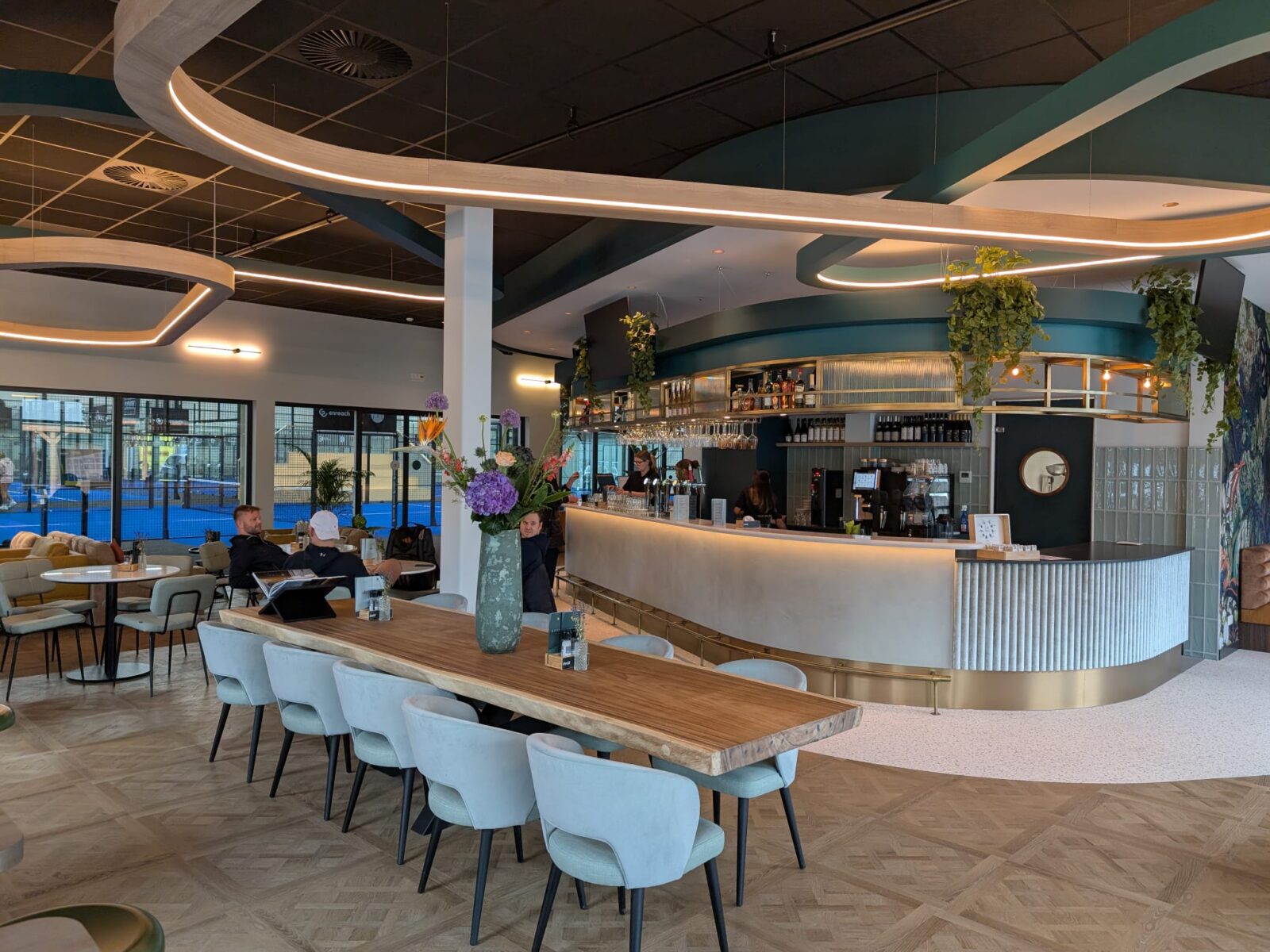 De horecazone van Poort Padel met een ronde bar, een lange houten tafel en uitzicht op de blauwe padelbanen