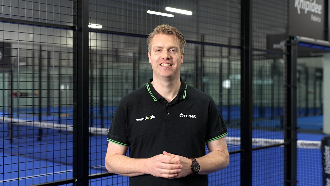 De presentator van de video in een zwart Eventlogic- en Reset-shirt voor een padelbaan.
